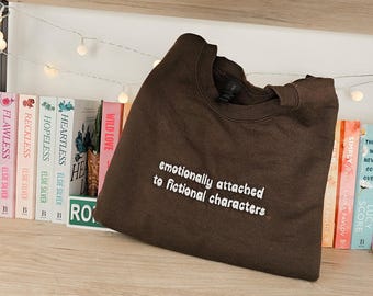 Op de afbeelding: Een donkerbruine sweatshirt met de witte tekst "emotionally attached to fictional characters". Het kledingstuk ligt opgevouwen op een houten plank, met boeken op de achtergrond.