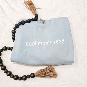 May include: A light blue sweatshirt with the text "cool moms read." in white. The sweatshirt is folded and placed on a white surface. A decorative beaded strand with a tassel is draped around the sweatshirt.