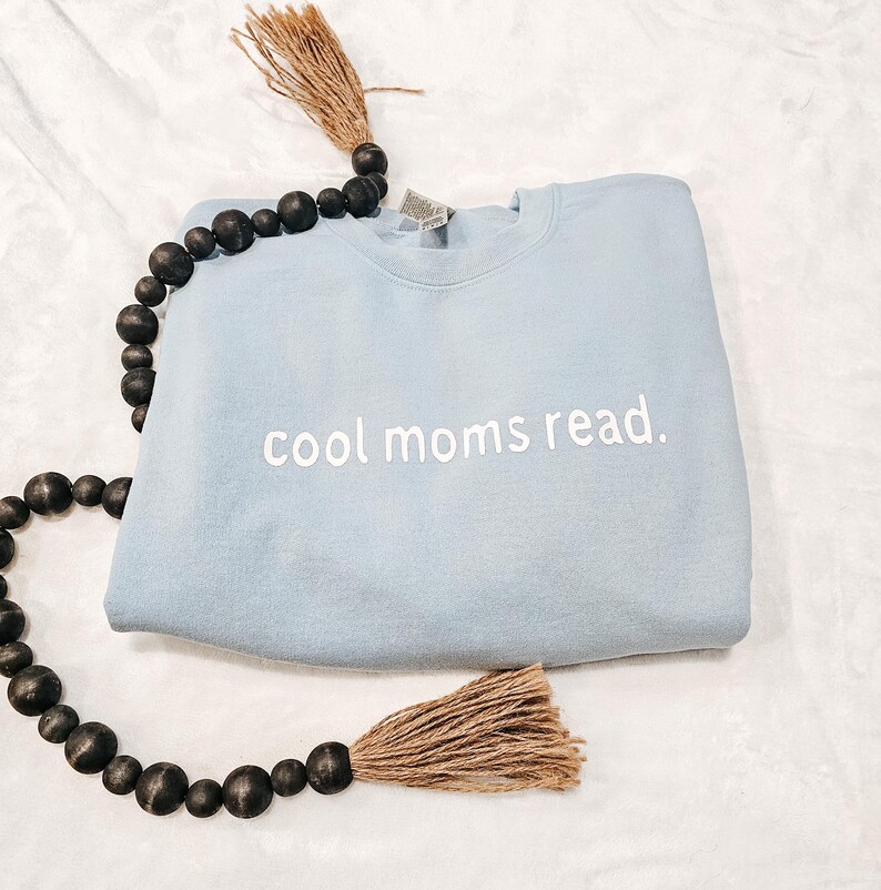 May include: A light blue sweatshirt with the text "cool moms read." in white. The sweatshirt is folded and placed on a white surface. A decorative beaded strand with a tassel is draped around the sweatshirt.