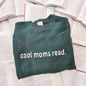 May include: A dark green sweatshirt with the text "cool moms read." in white, resting on a bed of open books. The sweatshirt has a crew neck and a relaxed fit, perfect for casual wear.