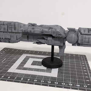 May include: A gray model of a spaceship, the Sulaco, is displayed on a black stand. The model has intricate details and is set against a white background with a cutting mat.