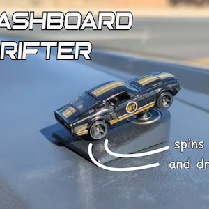 May include: A black and gold toy car with the text "Dashboard Drifter" and the words "spins and drifts!" The toy car has a racing stripe design and the number 67 on the side. The car is mounted on a black base.
