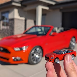 May include: A small red die-cast convertible toy car held in front of a full-sized red convertible car. The toy features a detailed front grille, silver-toned wheels, and an open interior, mirroring the design of the larger vehicle parked behind it.