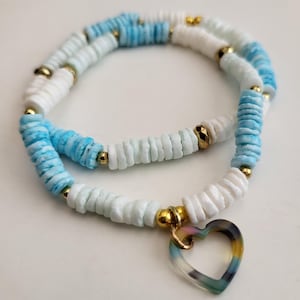 May include: A layered necklace featuring white and blue disc beads with gold accents. A heart-shaped pendant with a translucent, multicolored design hangs from the necklace. The necklace is displayed on a white surface.
