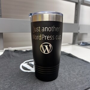 May include: Black stainless steel travel mug with a silver lid. The mug has the text "Just another WordPress cup" and the WordPress logo engraved on the front.