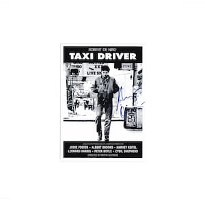 May include: Black and white movie poster for "Taxi Driver" featuring Robert De Niro. The poster includes the film's title, credits, and a signature. The image shows De Niro walking on a city street.