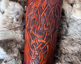 May include: A brown leather arm cuff with a Celtic knot design. The cuff has two metal clasps on the side.