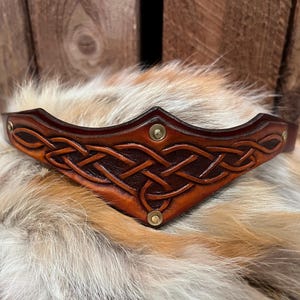 May include: A brown leather crown with a Celtic knot design. The crown is decorated with brass rivets and is set against a backdrop of fur and wood.