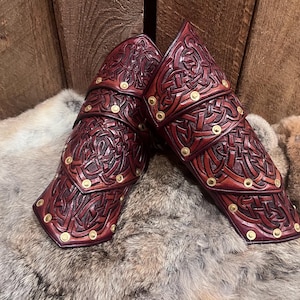 May include: Pair of burgundy leather arm guards with intricate Celtic knotwork and brass rivets. The arm guards are layered and designed to protect the forearm. They are resting on a bed of fur.