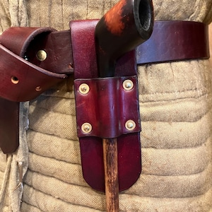 May include: A dark burgundy leather belt featuring a matching leather holster attached with four brass rivets. A wooden handle with a dark, polished, rounded top is inserted into the holster, which is secured around a textured beige garment.