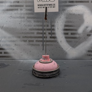 May include: A unique desk accessory made from a repurposed spray paint can. The base is pink and black, supporting a metal clip holding a white card with the text "MAG" repeated three times. The website address is also visible.