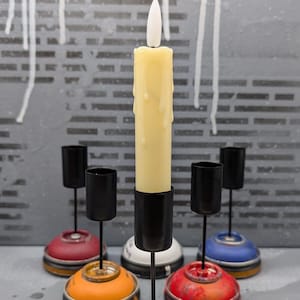May include: A collection of handmade candle holders. The candle holders are made from repurposed spray paint cans. The bases are various colors, including red, yellow, orange, and blue. A tall, cream-colored candle is in the center.