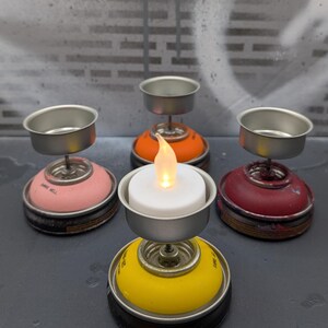 May include: A collection of handmade candle holders crafted from repurposed spray paint cans. The holders feature a variety of colors, including pink, orange, red, and yellow, each topped with a silver metal cup to hold a candle. One holder has a lit LED candle.