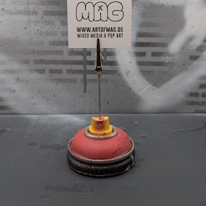 May include: A red and yellow spray paint can base holds a metal clip and a white card with the text "MAC MAC MAC" and "WWW.ARTOFMAG.DE MIXED MEDIA & POP ART". The background is a textured gray surface.