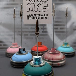 May include: A collection of six colorful spray paint can card holders. Each holder features a different color base, including pink, blue, red, and teal. A metal clip extends from each can, holding a white card with the text "MAG MAG MAG" and website address.