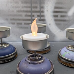 May include: A collection of handmade candle holders. Each holder is made from repurposed spray paint cans, featuring a small metal cup to hold a tea light candle. The candles have a warm, flickering glow.