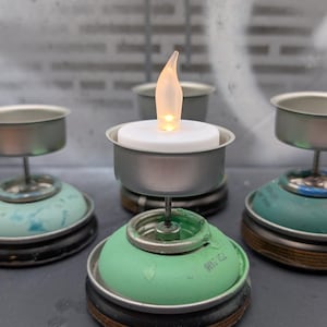 May include: A collection of handmade tealight candle holders. Each holder is crafted from repurposed materials, featuring a metal base and a small, flickering LED candle. The holders are in various shades of green and silver, creating a rustic, eco-friendly aesthetic.