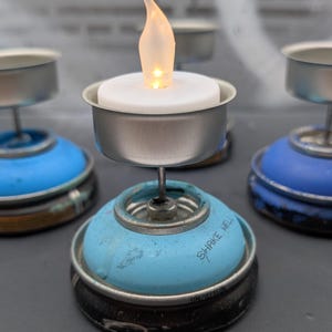 May include: A collection of tealight candle holders made from repurposed spray paint cans. Each holder features a small, battery-operated candle with a flickering flame. The bases are painted blue and black, with the words "SHAKE WELL" visible on one.