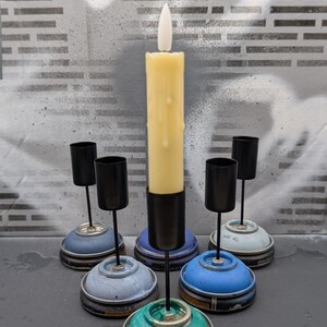 May include: A collection of handmade candle holders and a tall, ivory-colored candle. The candle holders are crafted from repurposed spray paint cans, featuring black metal cups and stems. The bases are painted in shades of blue and teal.