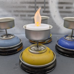 May include: A collection of handmade candle holders. Each holder is made from repurposed spray paint cans in yellow and blue, topped with a metal cup and a flickering LED candle. The background is a textured gray surface.