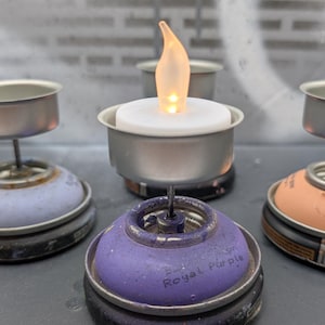 May include: A collection of handmade candle holders, each featuring a small LED candle with a flickering flame. The bases are repurposed spray paint cans in various colors, including purple and orange. The text "Royal Purple" is visible on one base.