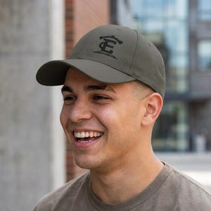 May include: Olive green baseball cap with a curved brim. The cap features a black embroidered design on the front. The cap is made of a durable fabric and has a classic six-panel design.
