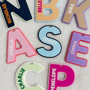 May include: A selection of brightly coloured letter-shaped name tags. Each tag displays a different name in white text, including "Nicholas," "Belle," "Kira," "Amelia," "Sonny," "Emma," "Charlie," and "Penelope." The tags are in various colours, such as pink, blue, and green.