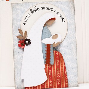 May include: A white card with a blue and white patterned background. A paper cutout of Mary holding baby Jesus is layered on top of the background. The cutout is surrounded by a white circle with the text "A little babe, so sweet & small".