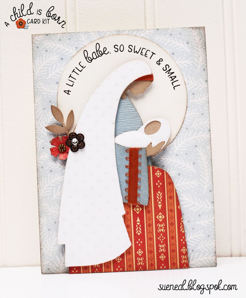 May include: A white card with a blue and white patterned background. A paper cutout of Mary holding baby Jesus is layered on top of the background. The cutout is surrounded by a white circle with the text "A little babe, so sweet & small".