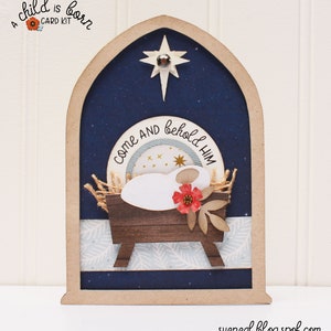 May include: A wooden Christmas card with a blue background and a star at the top. The card features a manger scene with a baby Jesus, a red flower, and the text "Come and behold Him".