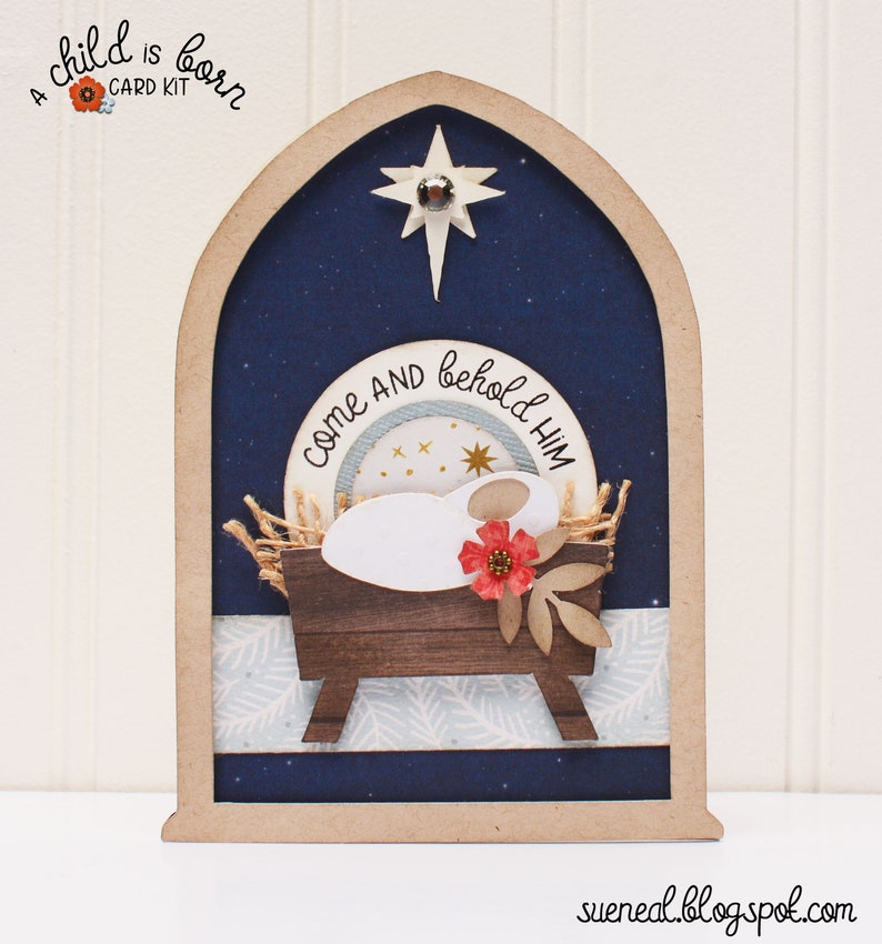 May include: A wooden Christmas card with a blue background and a star at the top. The card features a manger scene with a baby Jesus, a red flower, and the text "Come and behold Him".