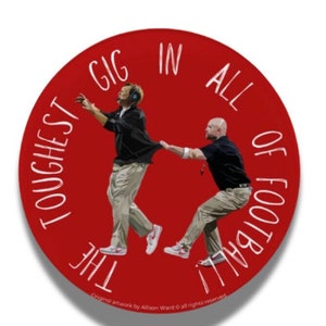 May include: A red circular button with white text that reads "The toughest gig in all of football". Two figures are depicted on the button, one in a black jacket and khaki pants, the other in a black shirt and khaki pants. The figure in the black jacket is holding his hands up in the air, while the figure in the black shirt is holding onto the other figure's jacket. The button is a Kirby Smart Game Day Button.