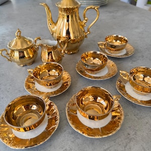 Puede incluir: Un juego de té dorado completo, que incluye una tetera, azucarero, lechera y varias tazas de té con platillos. El juego tiene una superficie brillante y reflectante y un diseño decorativo alrededor de los bordes de los platillos y las tazas. El juego está dispuesto sobre una superficie gris.