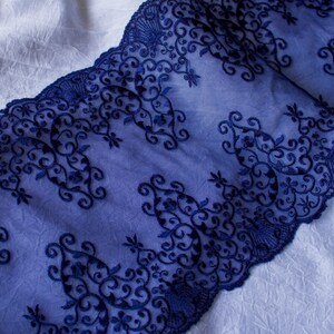 Puede incluir: Primer plano de tela de encaje azul marino con intrincados bordados florales y de volutas. El encaje est&aacute; superpuesto sobre una tela blanca texturizada, creando un contraste de color y textura. El dise&ntilde;o presenta un borde festoneado.