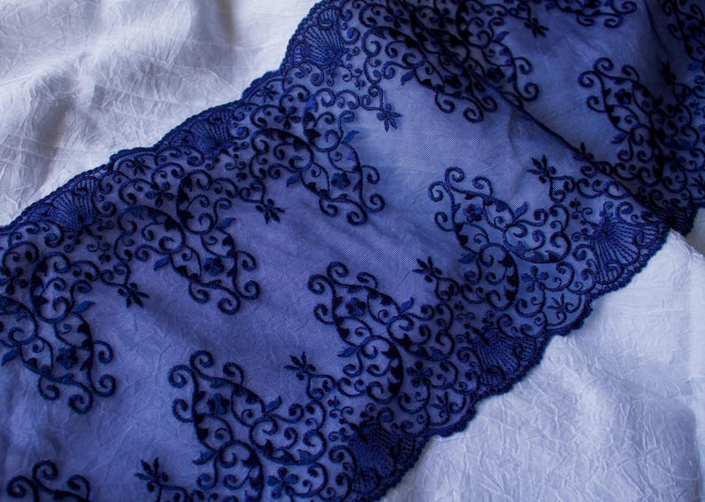 Puede incluir: Primer plano de tela de encaje azul marino con intrincados bordados florales y de volutas. El encaje est&aacute; superpuesto sobre una tela blanca texturizada, creando un contraste de color y textura. El dise&ntilde;o presenta un borde festoneado.