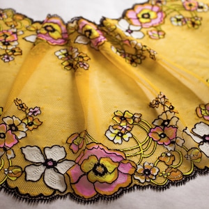 Op de afbeelding: Gele bloemenkant met zwarte gegolfde randen. De kant heeft roze, gele en witte bloemen met zwarte contouren.