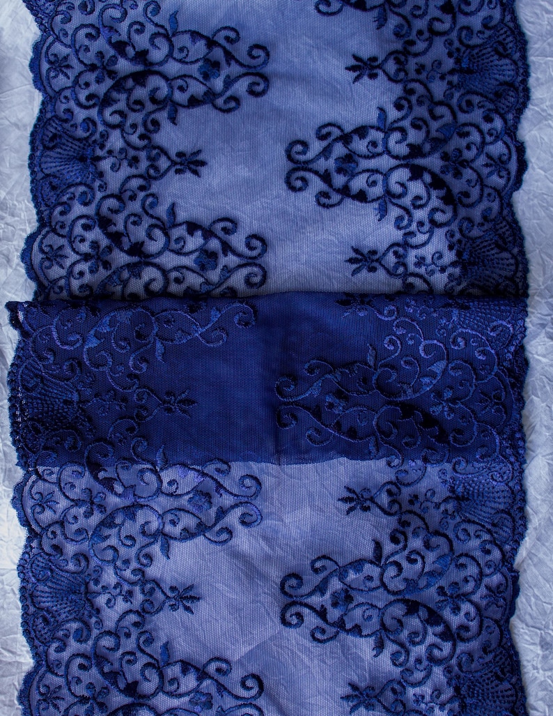 Puede incluir: Primer plano de una tela de encaje azul marino con intrincados patrones florales y de volutas. El encaje tiene un borde festoneado y est&aacute; superpuesto a una tela azul m&aacute;s clara, creando un contraste de color y textura. El dise&ntilde;o es sim&eacute;trico y detallado.