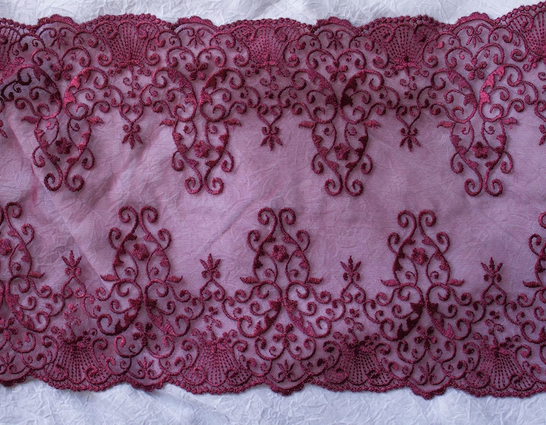 Puede incluir: Tela de encaje burdeos con un intrincado dise&ntilde;o floral y de volutas. El encaje presenta un borde festoneado y est&aacute; sobre un fondo lavanda suave. El bordado detallado crea una textura delicada y elegante, adecuada para diversos proyectos de manualidades.