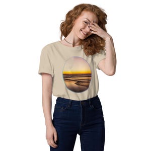 Könnte beinhalten: Beiges T-Shirt mit einem Sonnenuntergang-Motiv in ovaler Form. Das Design zeigt einen Strand mit Wellen, einen goldenen Sonnenuntergang und einen Himmel mit Farbverlauf. Das T-Shirt ist aus weichem Material und hat kurze Ärmel.