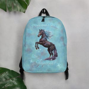 Puede incluir: Mochila azul claro con un caballo marrón encabritado, con melena y cola azules. El texto "Rise and Shine" y "Be Jupiter!" está escrito en la parte delantera. La mochila tiene correas negras y un asa superior, con estrellas.