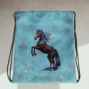 Puede incluir: Mochila con cordón con una ilustración de un caballo encabritado. El caballo es marrón oscuro con acentos azules y morados, sobre un fondo azul claro. El texto dice "Rise and Shine Be Jupiter!" en cursiva.