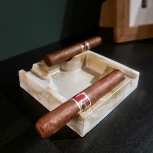 May include: A white ceramic ashtray with two brown cigars resting inside. The ashtray has a marbled design with gold accents.