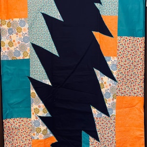 Könnte beinhalten: Ein Patchwork-Quilt mit einem marineblauen Blitzdesign. Der Quilt verwendet orangefarbene, türkisfarbene und florale Stoffe in Rot, Weiß und Blau. Der Artikel ist handgefertigt, ideal für ein Bett oder eine Dekoration.