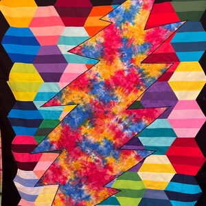 Könnte beinhalten: Ein farbenfrohes Quilt mit einem Batik-Blitzdesign auf einem Hintergrund aus mehrfarbigen sechseckigen Formen. Das Quilt zeigt eine breite Palette von Farben, darunter Rot, Blau, Gelb, Grün und Lila, wodurch ein visuell auffälliges Textil entsteht.