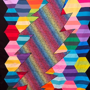 Könnte beinhalten: Ein farbenfrohes Quilt mit einem Regenbogen-Blitz-Design auf einem Hintergrund aus bunten sechseckigen Formen. Das Quilt besteht aus Stoff und hat einen schwarzen Hintergrund.