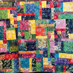 Op de afbeelding: Een kleurrijke patchworkquilt met een levendige mix van stoffen. De quilt heeft een verscheidenheid aan patronen en kleuren, waaronder rood, blauw, groen, geel en paars. Het ontwerp is een mix van vierkanten en rechthoeken.