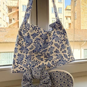 Peut inclure: Ensemble comprenant un sac fourre-tout, un chouchou et une trousse de maquillage. Les articles présentent un fond blanc avec un motif floral bleu. Le sac fourre-tout a une large bandoulière. Le chouchou est rond et gonflé. La trousse est en demi-lune.