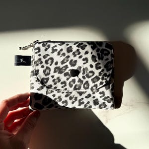 Peut inclure: Un petit portefeuille carré avec un motif léopard noir et blanc. Le portefeuille a une fermeture éclair, une fermeture à bouton-pression et une petite languette noire avec la lettre "K". Il est posé sur une surface blanche.
