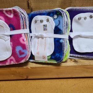 Puede incluir: Una pila de cuadrados de tela coloridos con inserciones blancas y broches. Los cuadrados de tela son de felpa y tienen diferentes patrones, incluyendo corazones, estrellas y diseños geométricos. Las inserciones son de algodón blanco y tienen broches en los bordes. Los cuadrados están atados juntos con una cinta blanca.