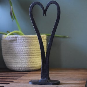 May include: A dark metal heart-shaped sculpture. The sculpture is made of a dark, possibly wrought iron, material. It stands upright on a small, flat base. The heart shape is formed by two curved lines that meet at the top. The sculpture is on a wooden surface.